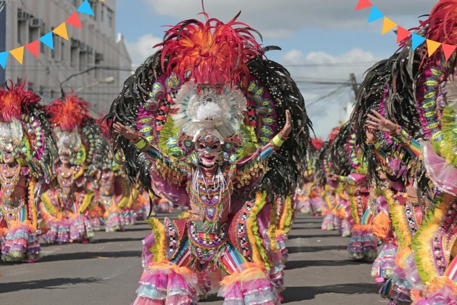 Dançarinos de samba em um desfile de Carnaval, com fantasias coloridas, plumas e carros alegóricos iluminados, representando a grandiosidade da festa brasileira.